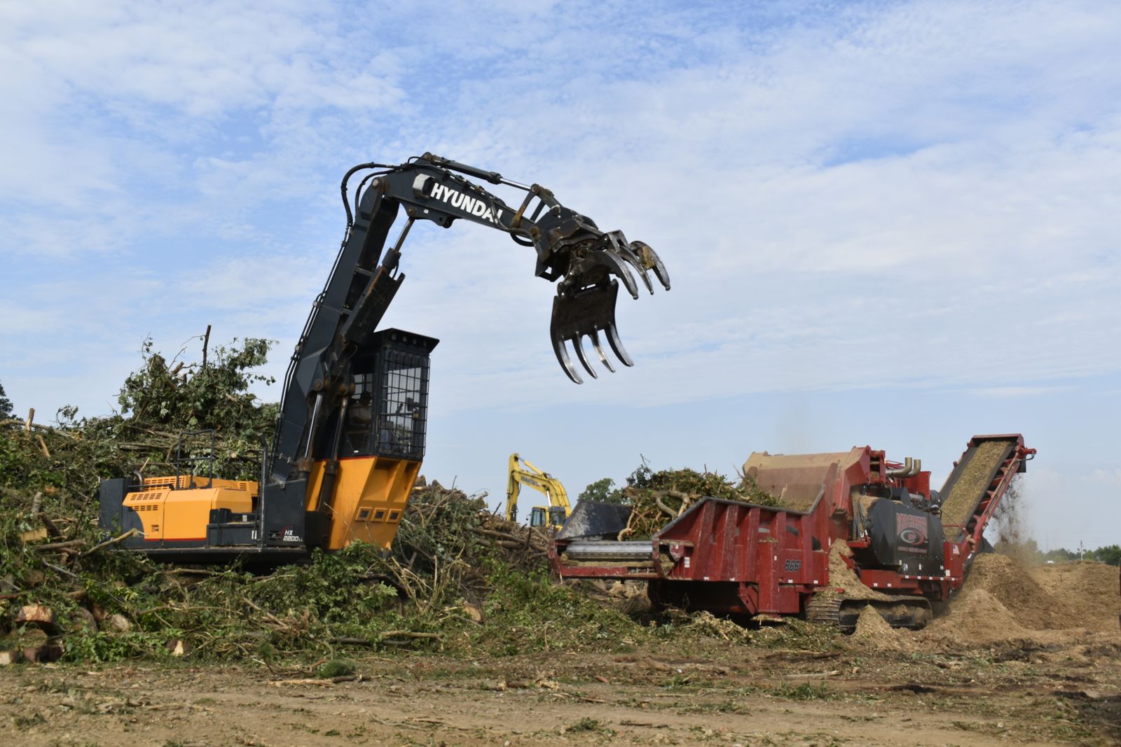 A grapple loader feeds tree limbs into a horizontal grinder at the EWP Mulch yard in Rogers, Arkansas, while freshly ground mulch pours off the discharge conveyor.