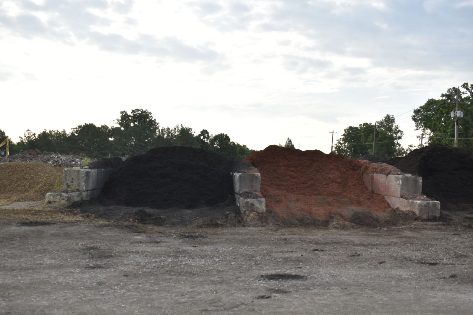 Concrete storage bays holding piles of black and red mulch at the EWP Mulch yard in Rogers, Arkansas, with wood-chip piles and trees in the background.